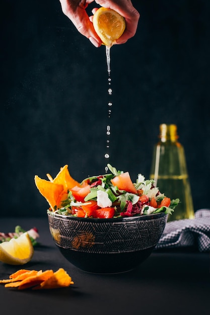 Salade de légumes dans un bol sur fond sombre avec une main féminine qui presse un citron sur un plat