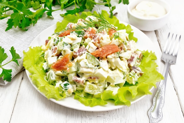 Salade d'oeufs de concombre de saumon et d'avocat habillés de mayonnaise sur des feuilles de laitue dans une assiette serviette aneth persil et fourchette sur fond de planche de bois blanc