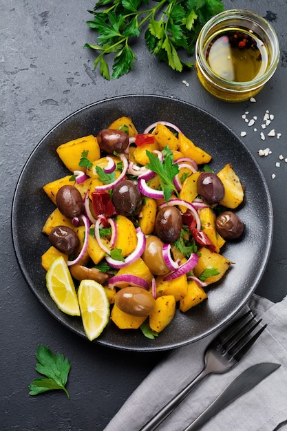 Salade de pommes de terre tiède aux olives, poivrons, persil et oignon rouge sur plaque en céramique noire sur fond de béton foncé