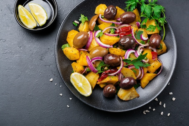 Salade de pommes de terre tiède aux olives, poivrons, persil et oignon rouge sur plaque en céramique noire sur fond de béton foncé