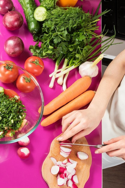 Salade préparée à la main féminine coupant des radis sur une table de cuisine rose