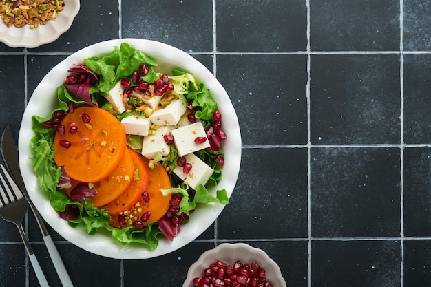 Salade Salade verte fraîche avec fromage feta kaki grenade et pistache sur une assiette blanche sur de vieux carreaux noirs Idée pour une délicieuse salade de Noël d'hiver saine Alimentation saine et équilibrée Vue de dessus