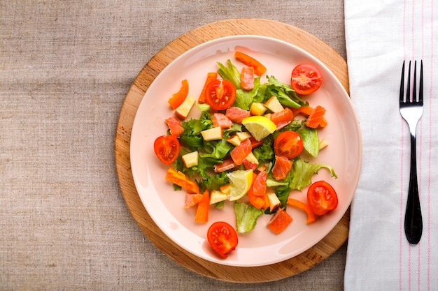 Salade de saumon et tomates cerises et salade verte dans une assiette sur une table en bois sur un support en bois avec une fourchette et une serviette à côté.