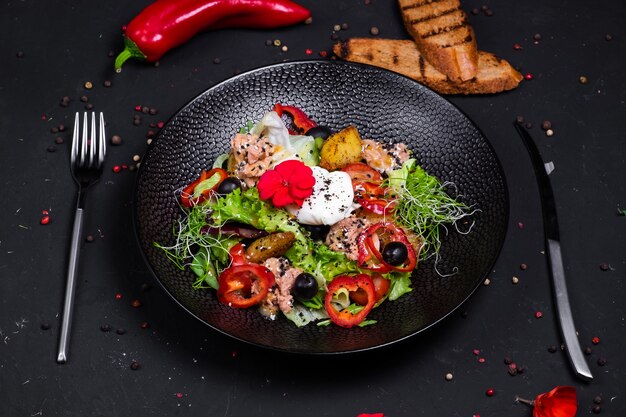 Salade de thon en combinaison avec pommes de terre frites, poivrons, olives, œuf poché, sur un oreiller avec un mélange de salade, et une vinaigrette à base de deux moutardes, sur une surface sombre