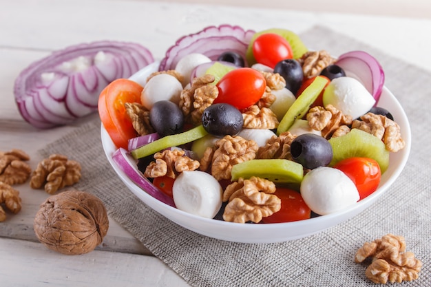 Salade de tomates cerises, fromage mozzarella, olives, kiwi et noix sur une surface en bois blanche.