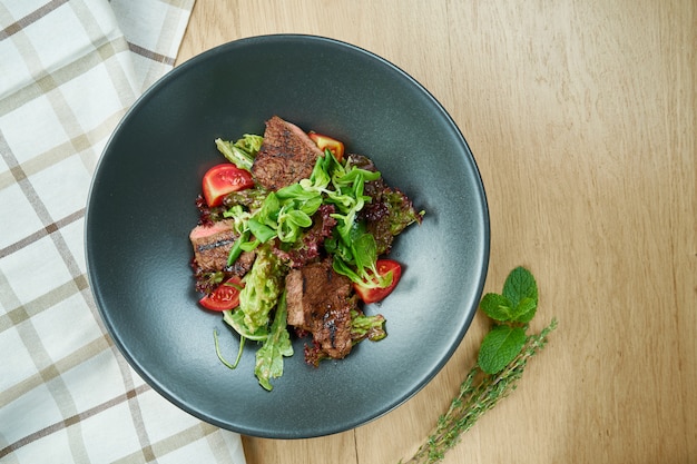Salade de veau grillée avec laitue, tomates cerises, fromage bleu dor dans un bol noir sur table en bois. Mise au point sélective, gros plan