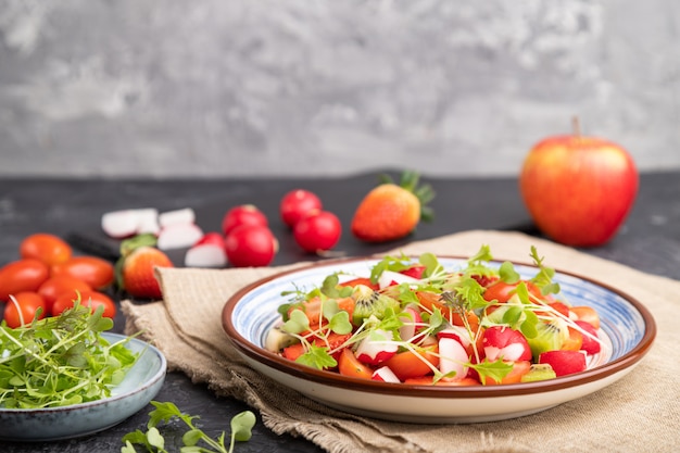 Salade végétarienne de fruits et légumes de fraise, kiwi, tomates, pousses microgreen sur fond noir et gris et textile en lin.