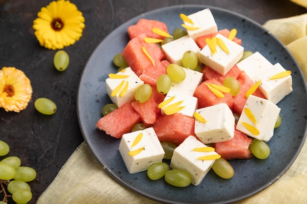 Salade végétarienne avec pastèque, fromage feta et raisins sur plaque en céramique bleue sur fond de béton noir et textile en lin jaune. Vue latérale, gros plan, mise au point sélective.