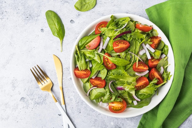 Salade verte d'été dans une assiette de bol blanche sur une table grise. Salade saine de tomates, épinards, oignons et feuilles vertes avec de l'huile pour le dîner ou le déjeuner. Concept de cuisine végétarienne. Vue de dessus sur fond de pierre grise