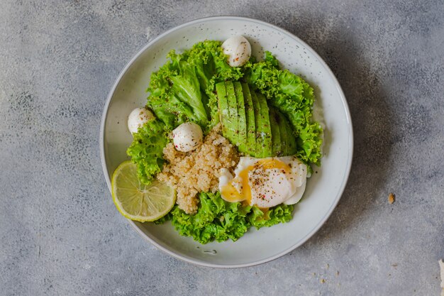 Saladier vert avec quinoa, mozzarella, avocat, œuf poché, laitue, citron vert et huile d'olive saisis sur une surface en pierre grise. concept de saine alimentation. fermer