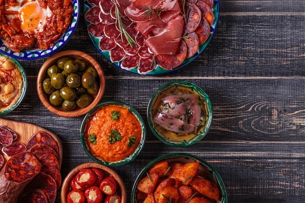 salami, bols aux olives, poivrons, anchois, pommes de terre épicées, purée de pois chiches sur une table en bois.