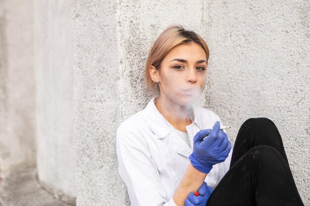 Sale infirmière fatiguée avec de la cendre sur le visage assise à l'extérieur de l'hôpital et fumant