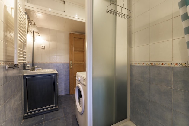 Salle de bain avec lavabo en porcelaine blanche avec dessus en marbre et meuble en bois bleu avec miroir intégré et appliques de la même couleur que le carrelage avec machine à laver et cabine de douche