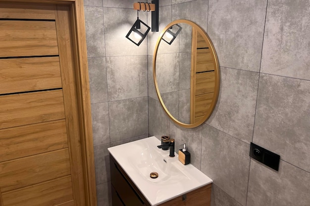 Une salle de bain spacieuse et élégante dans un thème bois et gris La salle de bain est dotée d'une porte en bois et d'un miroir ovale Le lavabo et le robinet sont blancs et contrastent avec le carrelage gris