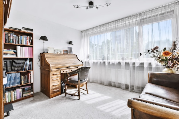 Salle blanche éclairée par le soleil avec secrétaire en bois et fauteuil près de la bibliothèque et de la fenêtre avec rideau blanc