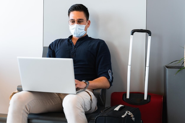 Salon d'attente pour homme à l'aéroport ou à la gare à côté de nombreux bagages Homme avec masque facial à l'aide d'un ordinateur portable