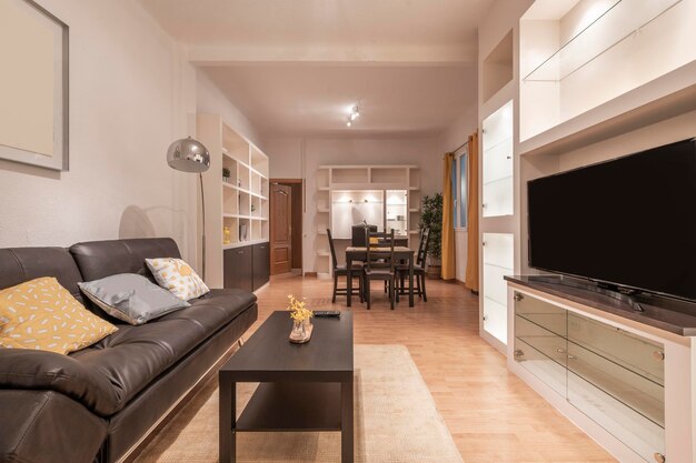 Salon avec canapé en cuir avec table basse assortie et bibliothèque blanche avec portes vitrées et télévision à écran plat parquet et table à manger en arrière-plan