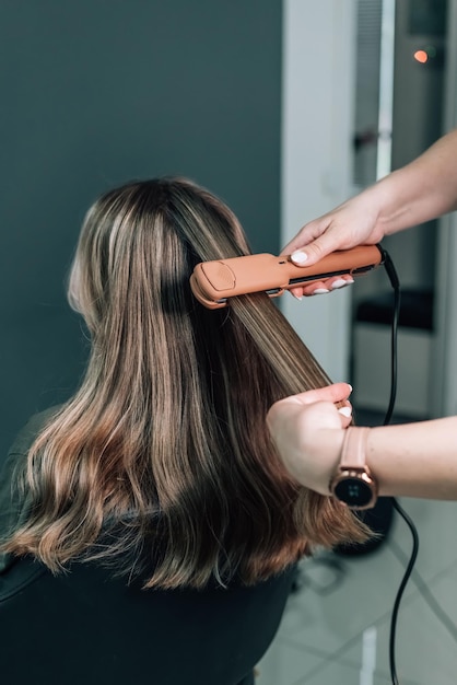 Salon de coiffure femme redresse les cheveux bruns d'une belle femme à l'aide de pinces à cheveux dans un salon de beauté styliste femme fait une coiffure pour une belle jeune femme dans un salon de coiffure