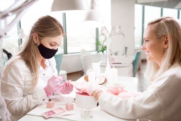 Salon de manucure et pédicure, covid-19 et distanciation sociale. Maître en gants en caoutchouc et jeune femme cliente en masque de protection à l'intérieur du studio de beauté