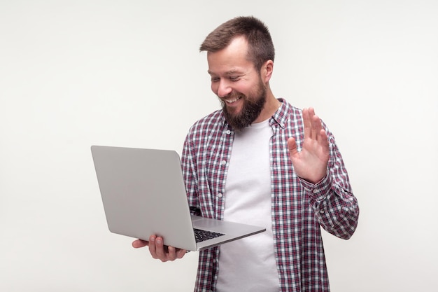 Salut. Portrait d'un homme barbu positif en chemise à carreaux décontractée agitant la main sur un ordinateur portable, faisant un appel vidéo, parlant en ligne sur le réseau social, visage souriant heureux. studio intérieur tourné isolé, fond blanc