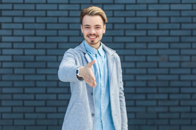 Salutation et poignée de main Portrait d'heureux beau jeune homme blond dans un style décontracté debout sourire à pleines dents et regardant la caméra et donnant la main pour secouer le studio intérieur tourné sur fond de mur de briques