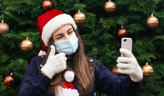 Salutations de Noël en ligne. Gros plan Portrait de femme portant un chapeau de père Noël et un masque médical avec émotion. Dans le contexte d'un arbre de Noël. Pandémie de Coronavirus