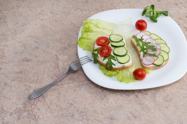 Sandwich au fromage cottage, concombres et tomates cerises et radis sur une plaque blanche sur la table. Assiette avec une fourchette sur la table avec une assiette blanche avec une salade de légumes.