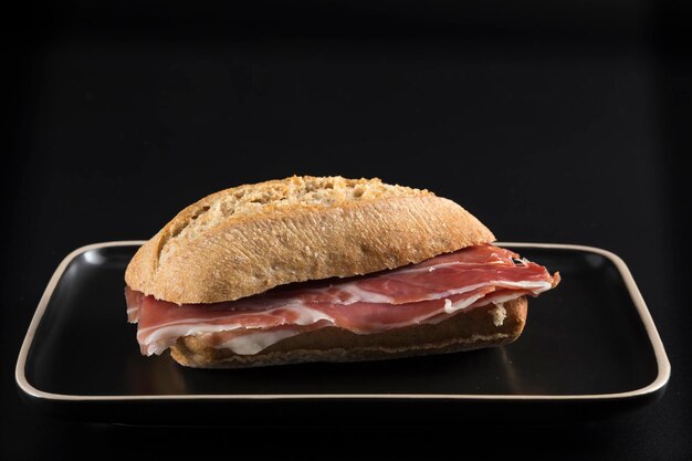 Sandwich au jambon ibérique sur un plateau de couleur foncée tout souligné par une bordure claire sur un fond noir