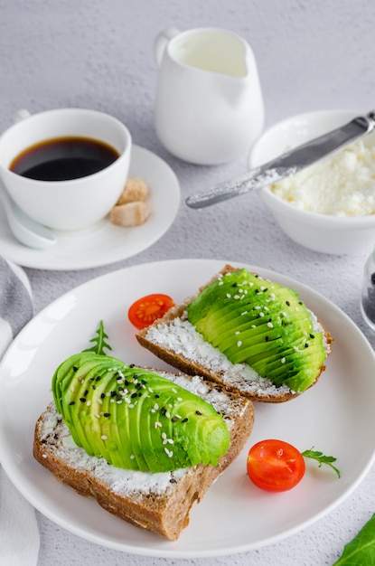 Sandwich à l'avocat sur pain de seigle noir fait avec de l'avocat en tranches fraîches, du fromage à la crème et des graines sur une plaque blanche avec une tasse de café sur un fond clair.