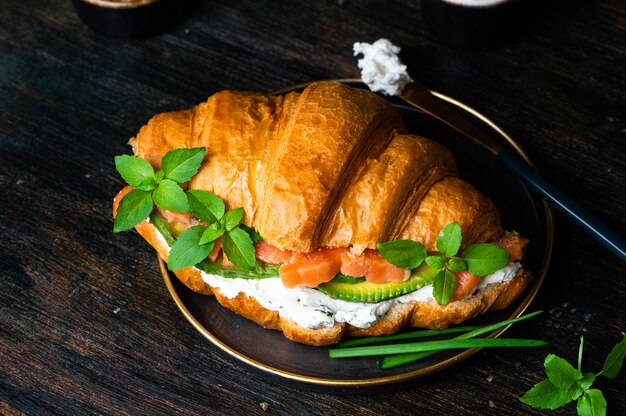 Photo sandwich croissant au saumon salé sur plaque noire, servi avec des feuilles de basilic frais, de l'avocat et du fromage philadelphia. petit déjeuner français. concept d'alimentation saine.