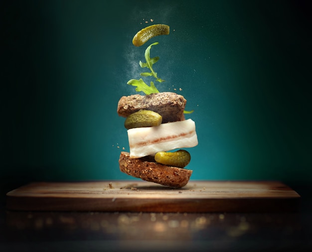 Photo un sandwich dans un style folk avec une tranche de bacon sur fond bleu avec des ingrédients volants cornichons et roquette