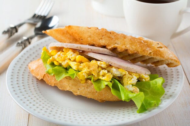 Sandwich farci aux œufs brouillés, au jambon et aux feuilles de laitue sur un fond en bois clair. Mise au point sélective.