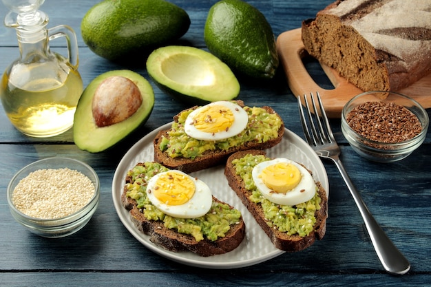 Sandwich avec purée d'avocat et œuf sur une assiette et ingrédients pour la cuisson sur une table en bois bleue.