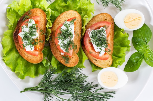 Un sandwich à la tomate, au fromage et aux légumes verts dans une assiette blanche.