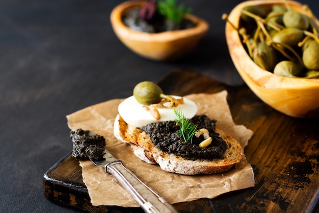 Sandwich avec tranche de fromage mozzarella et tapenade, câpres sur fond de table rustique sombre. Plat traditionnel provençal. Mise au point sélective