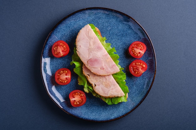 Sandwich à la viande de jambon de dinde, salade verte et tranches de tomates cerises fraîches sur une plaque brillante bleue, mur minimal bleu, vue de dessus