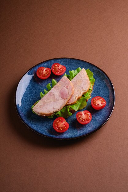 Sandwich à la viande de jambon de dinde, salade verte et tranches de tomates cerises fraîches sur une plaque brillante bleue, mur minimal brun, angle de vue