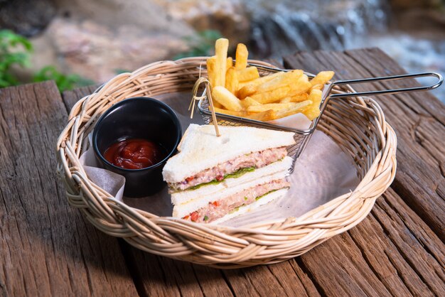 Sandwiches au thon servis avec croustilles et ketchup, disposés dans un beau panier en rotin, posé sur une table en bois.