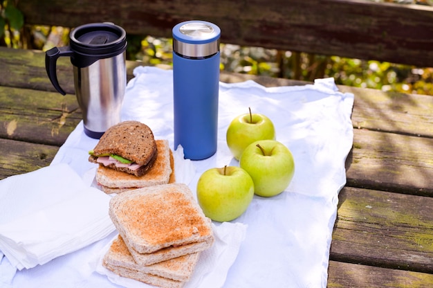 Photo sandwiches et deux thermos avec thé et café sur une table en bois dans le parc sur un fond de feuilles vertes, un arbre et la lumière du soleil. concept de tourisme et de voyage. espace copie