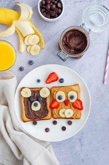 Sandwiches Drôles De Visage Sain Pour Les Enfants. Des Visages D'animaux Grillé Avec Beurre D'arachide Et Chocolat Noisette, Banane, Fraise Et Myrtille Sur Une Plaque Blanche Avec Du Jus D'orange.