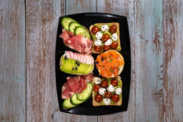 Sandwichs assortis avec du poisson, du fromage, de la viande et des légumes sur une plaque noire et un fond en bois