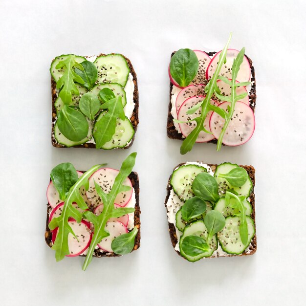 Sandwichs au fromage à la crème, concombre frais, radis, graines de chia et roquette sur papier blanc. Coups de nourriture au-dessus de la tête. Mise à plat