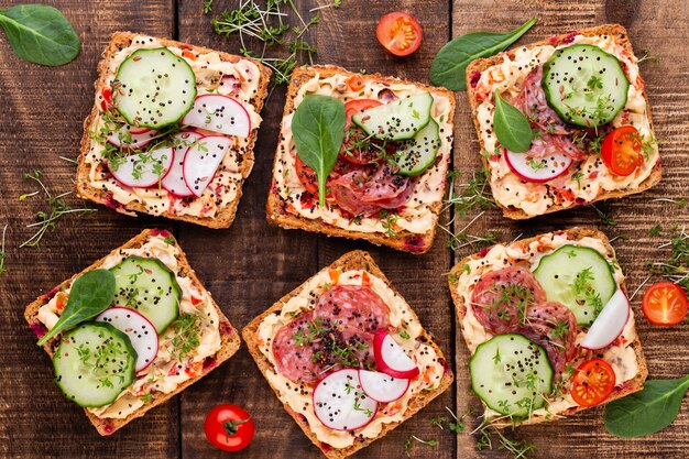 Sandwichs au fromage à la crème, légumes et salami. Sandwichs au concombre, radis, tomates, salami sur fond gris, vue du dessus. Mise à plat.