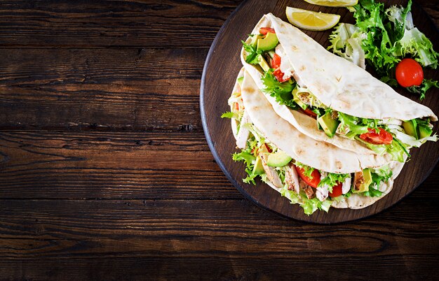 Sandwichs au pain pita avec viande de poulet grillée, avocat, tomate, concombre et laitue servis sur fond de bois.