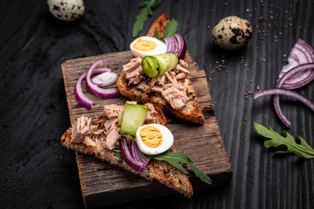 Sandwichs italiens à la bruschetta avec des œufs de caille et des oignons de concombre de thon en conserve Toasts au thon Délicieux petit-déjeuner ou collation sur fond sombre vue de dessus