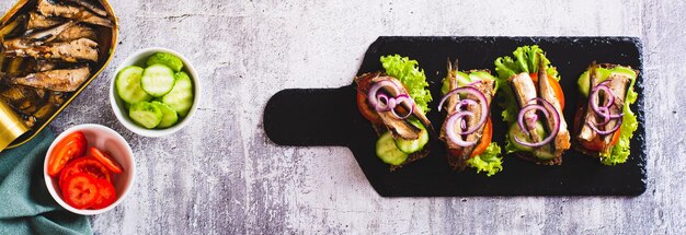 Sandwichs sur pain de seigle avec des sprats fumés des légumes et des herbes sur l'ardoise vue supérieure bannière web