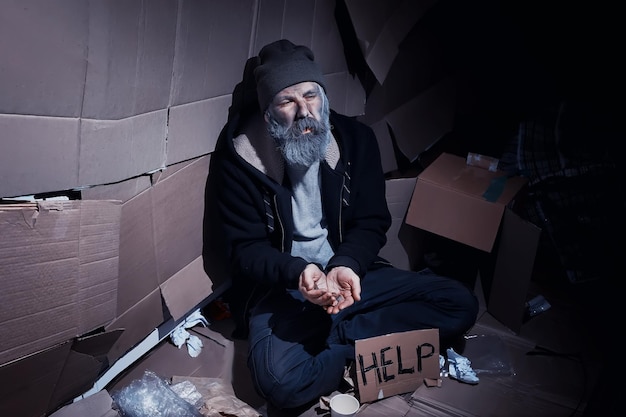 Un sans-abri barbu est assis sur des boîtes dans la rue et demande de l'aide Besoin d'un sans-abri demande de l'argent pour la nourriture et la nuit