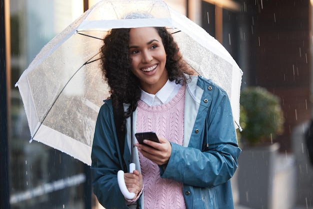 Sans pluie, il n'y a pas de fleurs. Photo d'une jeune femme utilisant un téléphone tout en tenant un parapluie dans la ville.