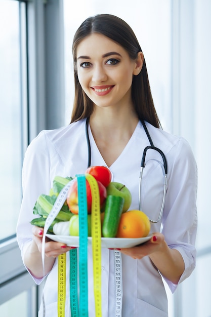 Santé et beauté. Un docteur sérieux écrit un régime d'avion. Femme assise au bureau. Jeune médecin avec beau sourire et fruits frais.