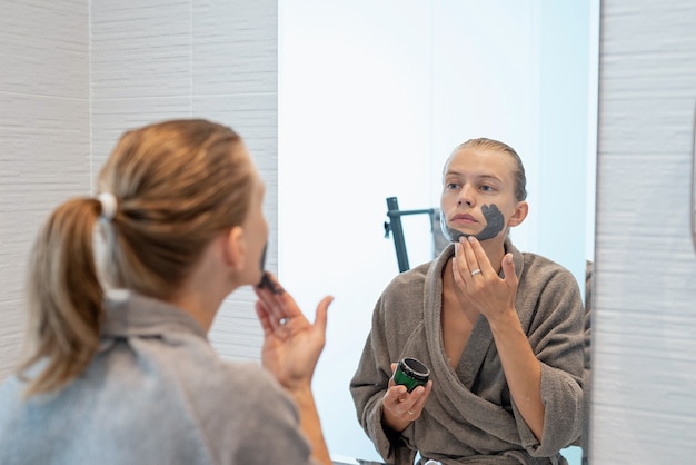 Santé des femmes. Spa et bien-être. Femme en peignoirs gris appliquant un masque facial dans la salle de bain en regardant le miroir